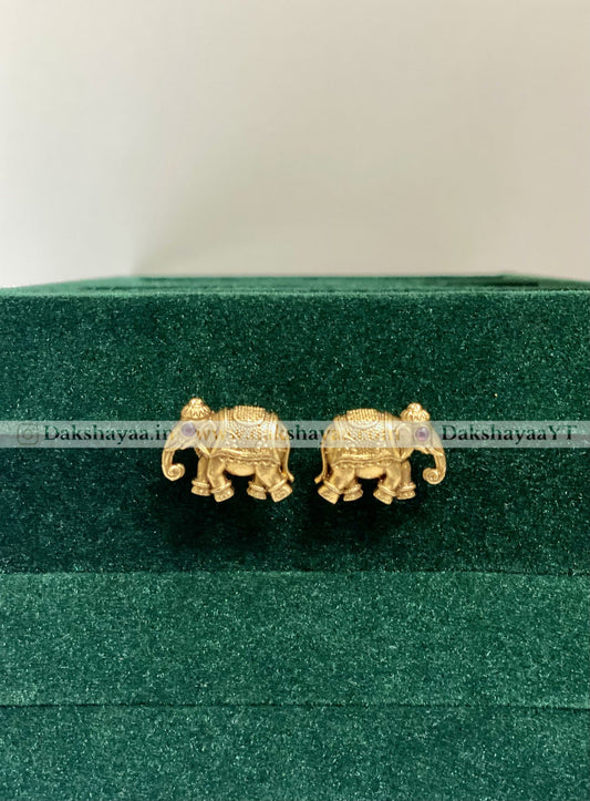 Gold elephant earrings on a green velvet cushion with 'Dakshayaa' branding.