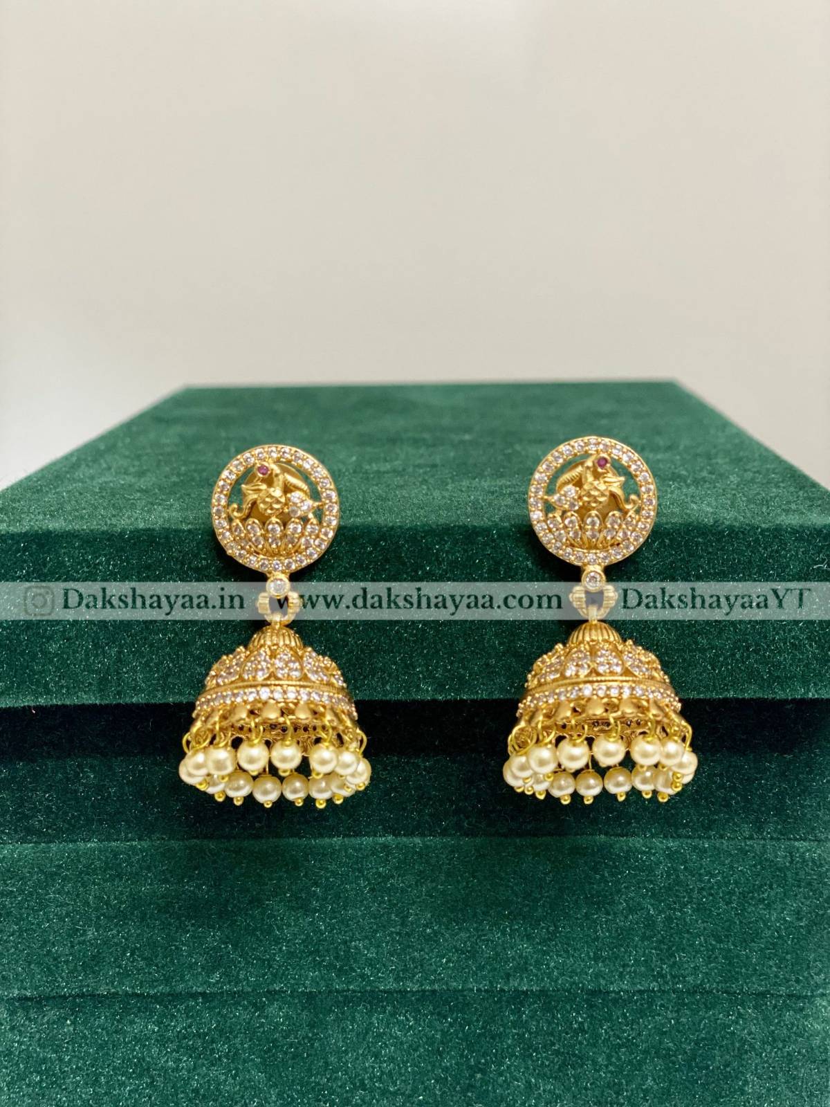 Gold jhumka earrings on a green velvet surface with 'Dakshayaa' branding.