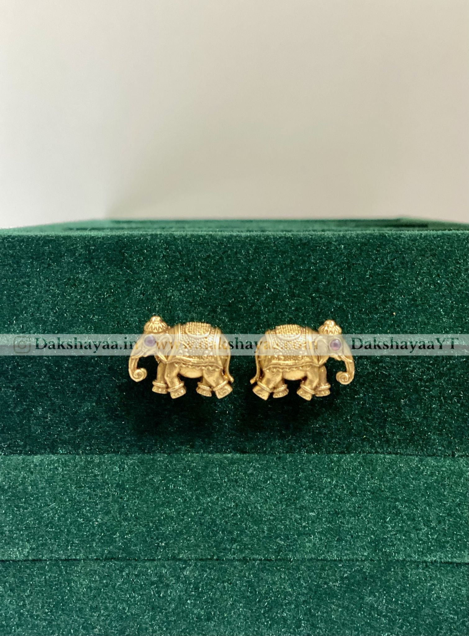 Gold elephant earrings on a green velvet cushion with 'Dakshayaa' branding.