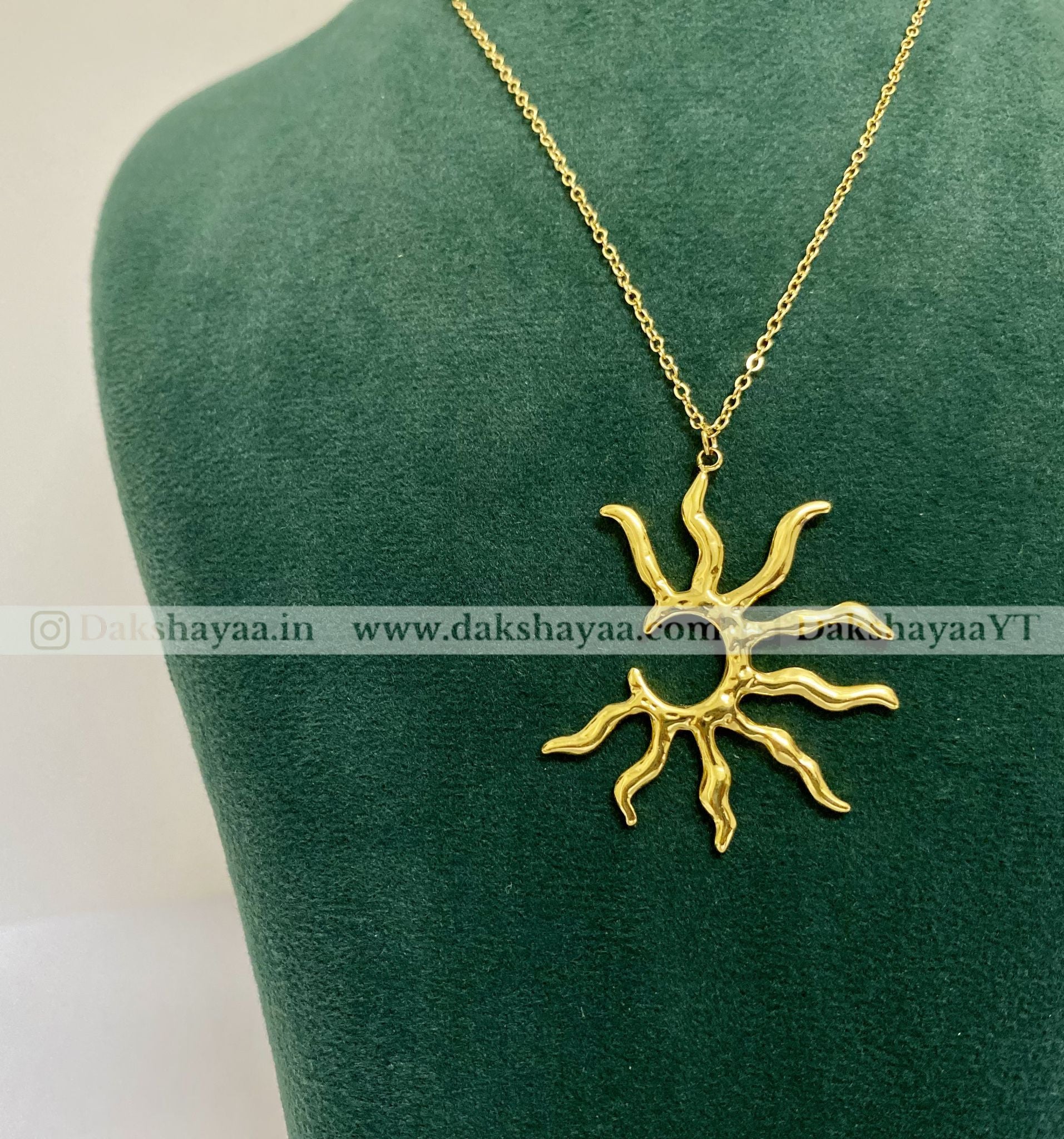 Gold sun-shaped pendant on a green mannequin with dakshayaa visible brand watermark.