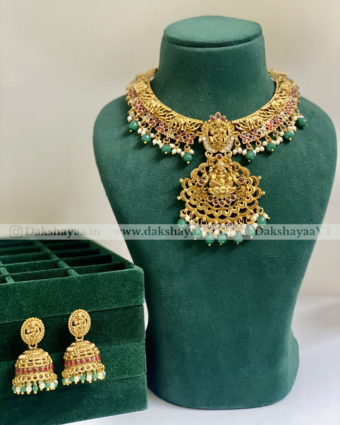Gold necklace and earrings on a green mannequin with 'Dakshayaa' branding.