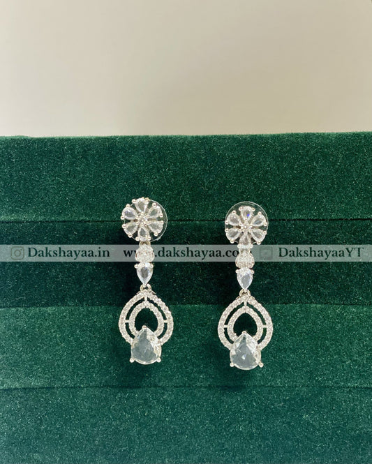 Silver earrings on a green velvet background with 'Dakshayaa.in' branding.
