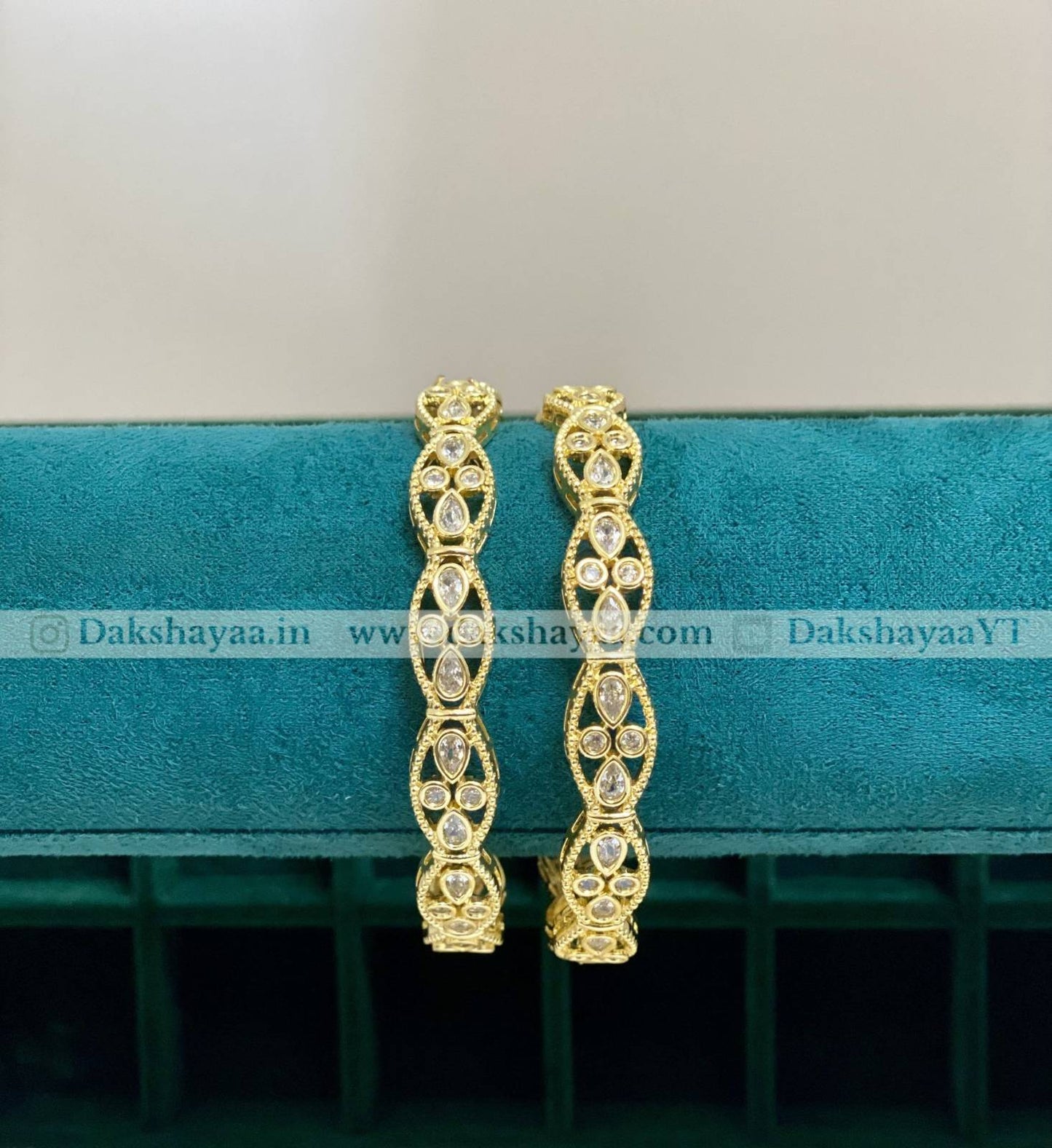Gold hoop bangles on a teal cushion with 'Dakshayaa.in' branding.