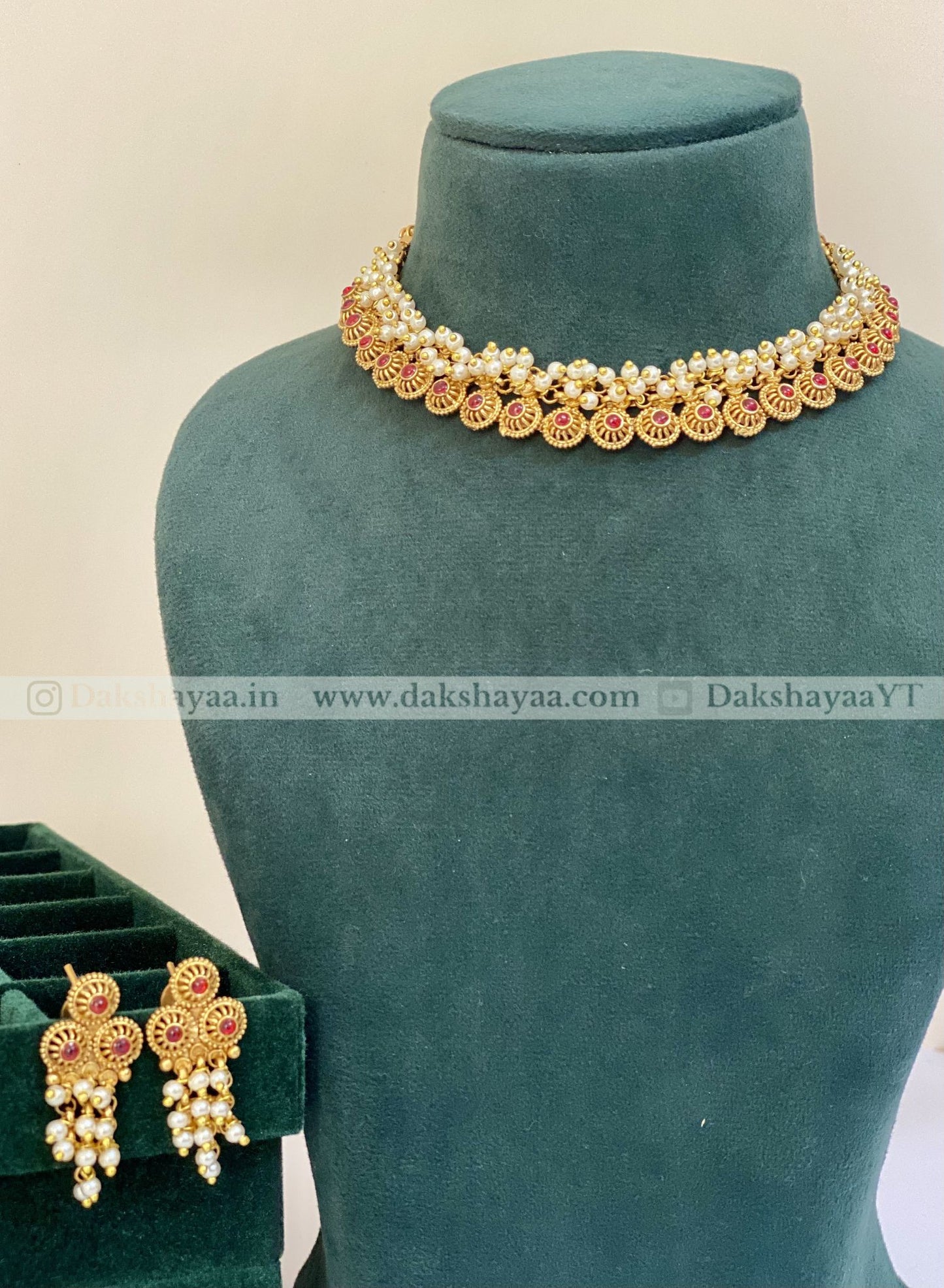 Gold necklace and earrings on a green velvet stand with 'Dakshayaa' branding.