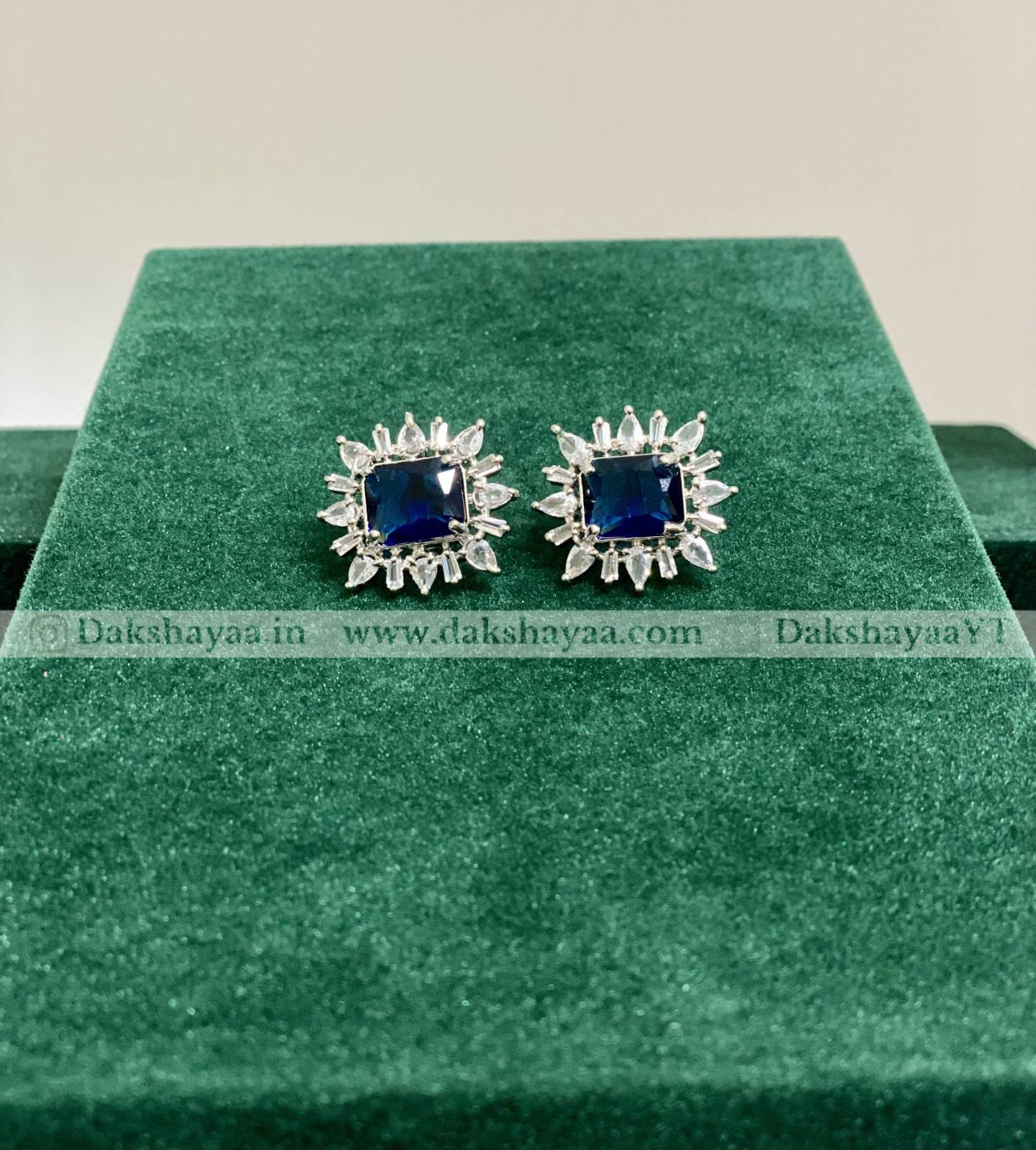 Pair of blue gemstone earrings on a green cushion with 'Dakshayaa' branding.