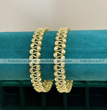 Gold bracelets with a wavy design on a teal cushion, brand 'Dakshayaa' visible.