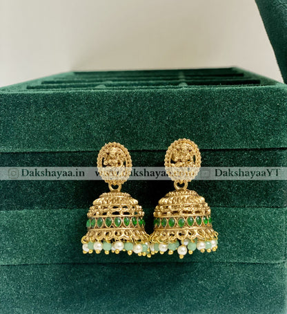 Gold and green earrings on a green velvet surface with 'Dakshayaa.in' branding.