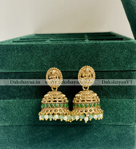 Gold and green earrings on a green velvet surface with 'Dakshayaa.in' branding.