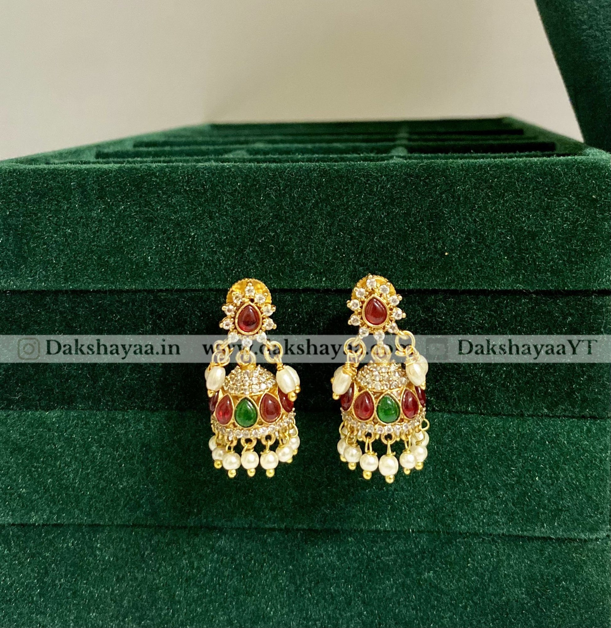 Ornate earrings with red and green stones on a green velvet surface, branded 'Dakshayaa'.