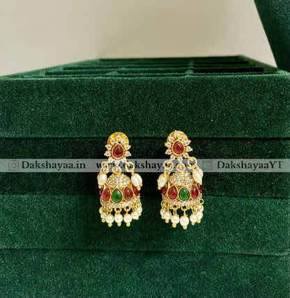 Ornate earrings with red and green stones on a green velvet surface, branded 'Dakshayaa'.