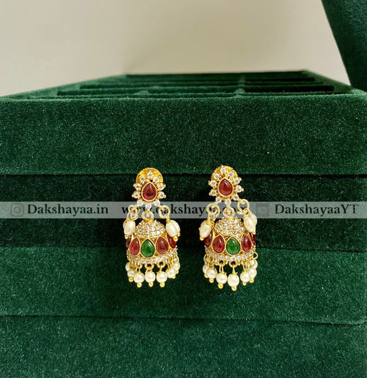 Ornate earrings with red and green stones on a green velvet surface, branded 'Dakshayaa'.