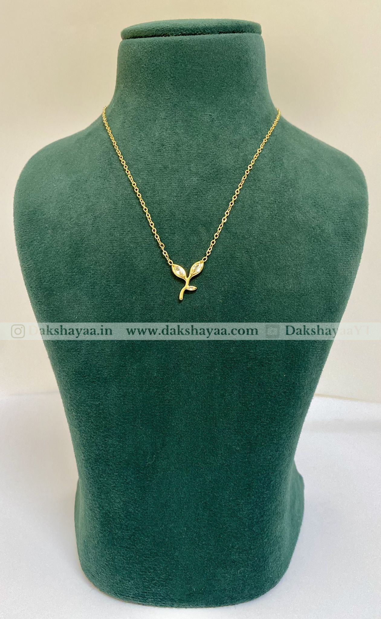 Gold necklace with a leaf pendant on a green mannequin bust, brand 'Dakshayaa' visible.