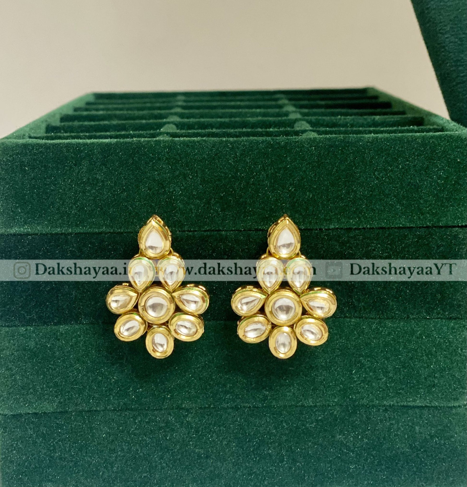 Gold and pearl earrings on a green velvet stand with 'Dakshayaa' branding.