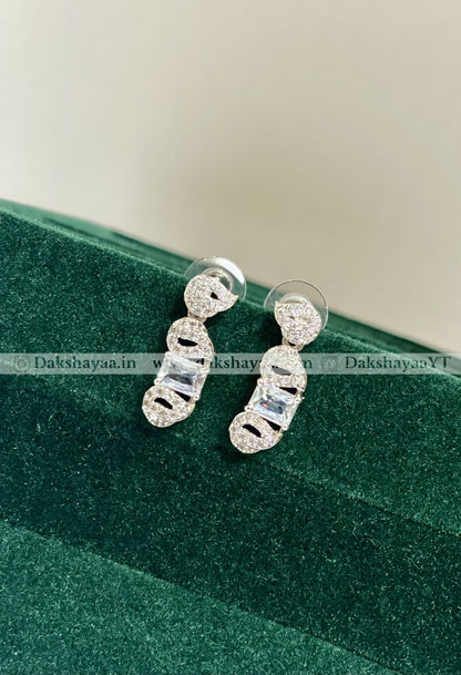 Silver earrings on a green textured surface with 'Dakshayaa.in' branding.