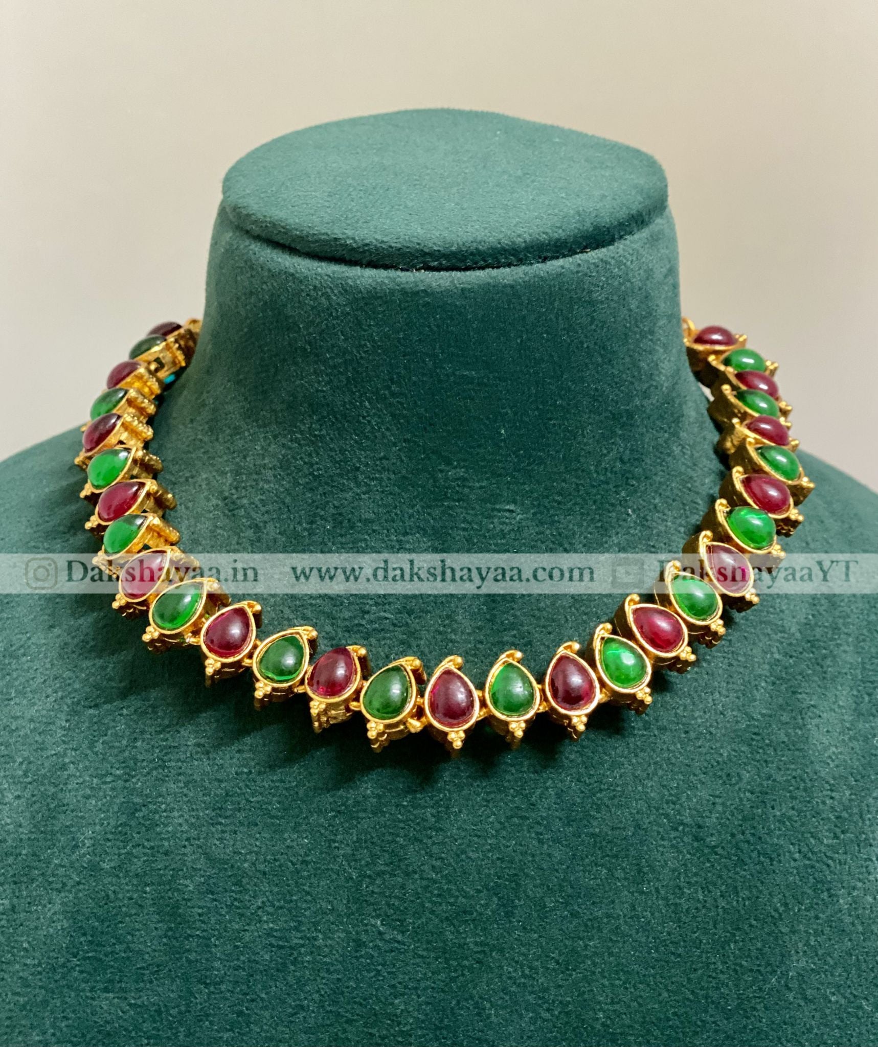 Gold necklace with green and red stones on a green velvet stand
