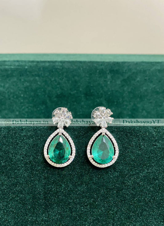 Teardrop-shaped emerald earrings on a green background with 'Dakshayana' branding.