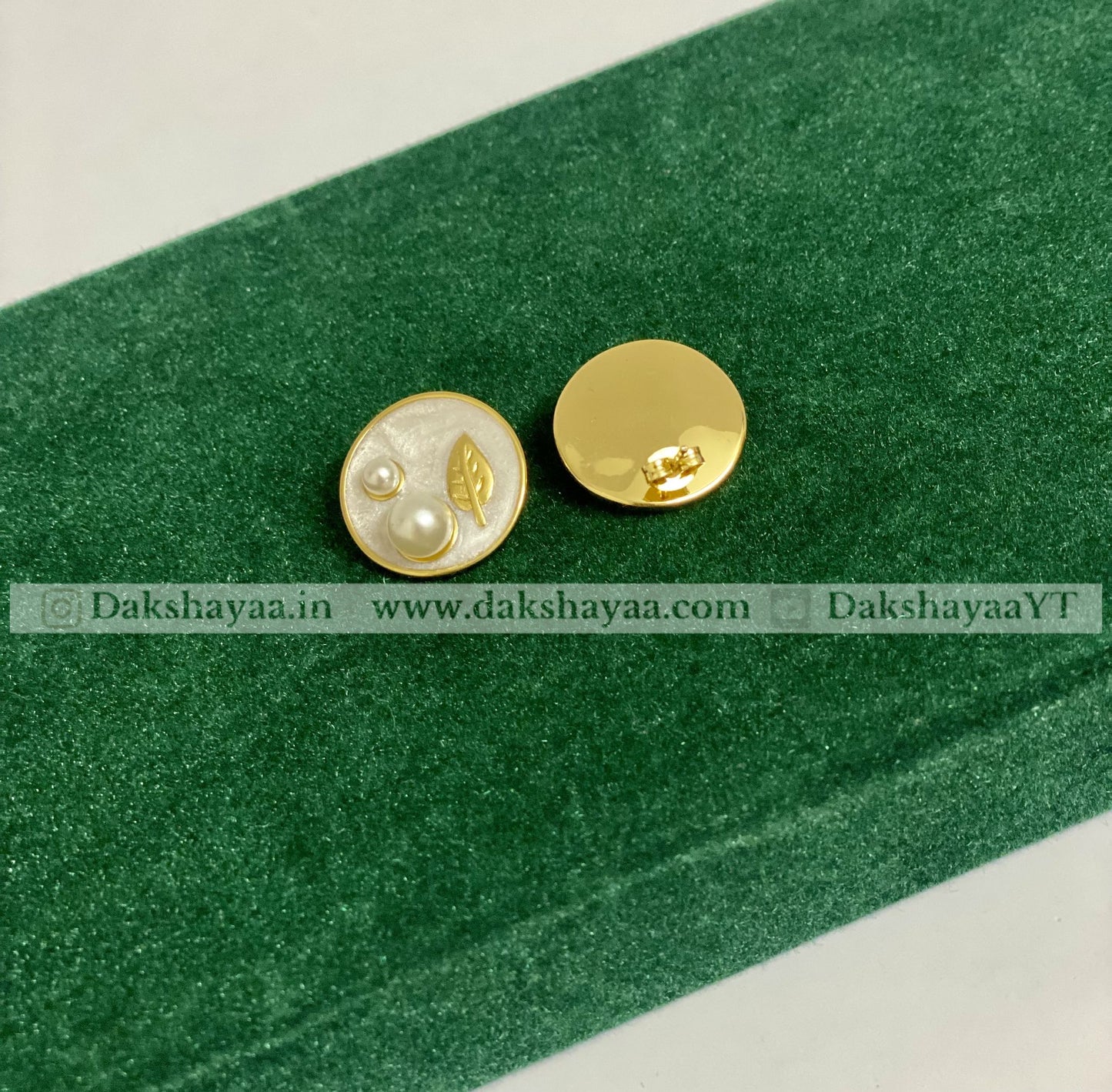 Pair of gold and white earrings on a green surface with dakshayaa branding text.