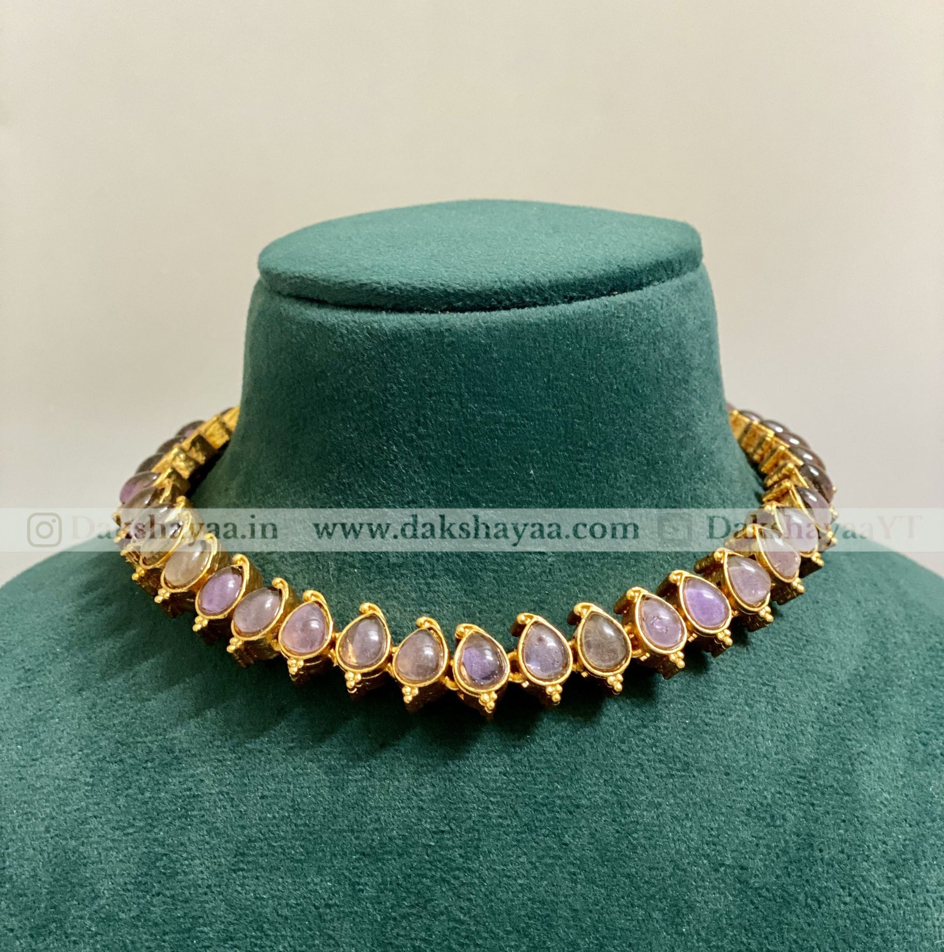 Gold necklace with purple stones on a green velvet stand, brand 'Dakshayaa' visible.