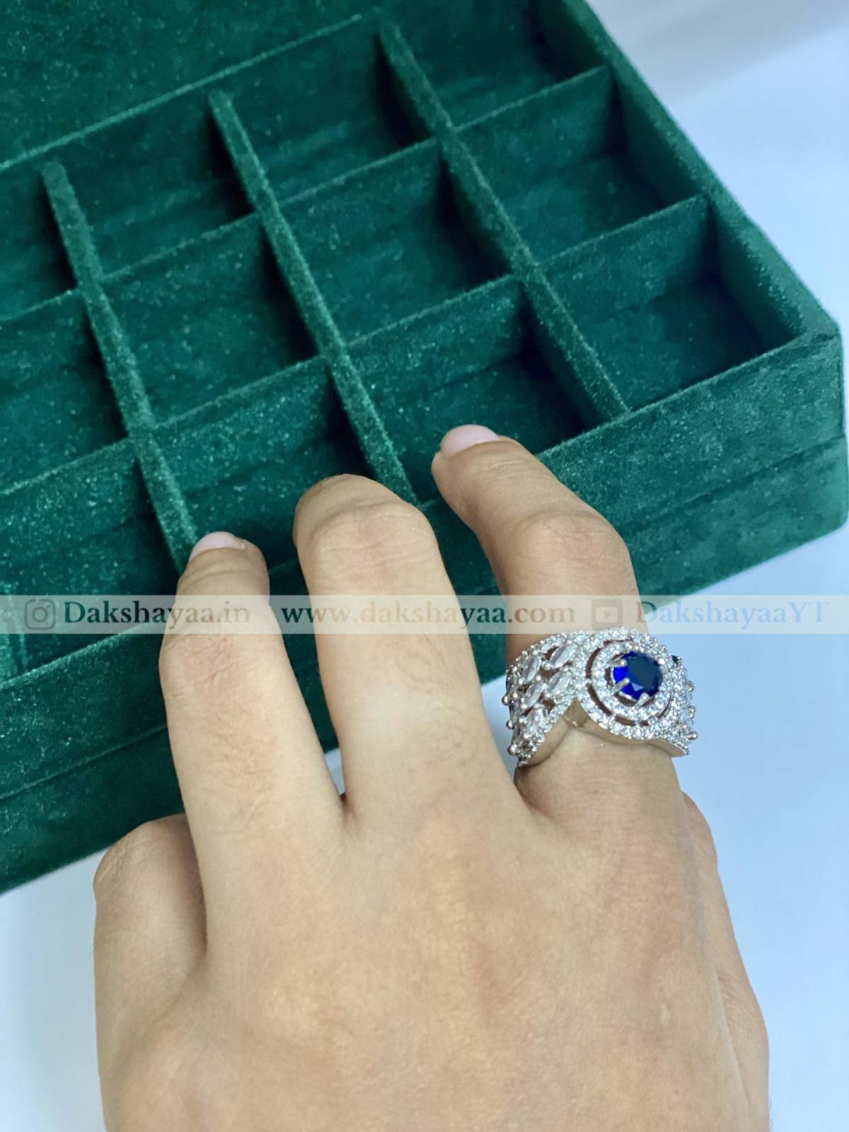 Hand wearing a ring with a blue gemstone against a green jewelry box background