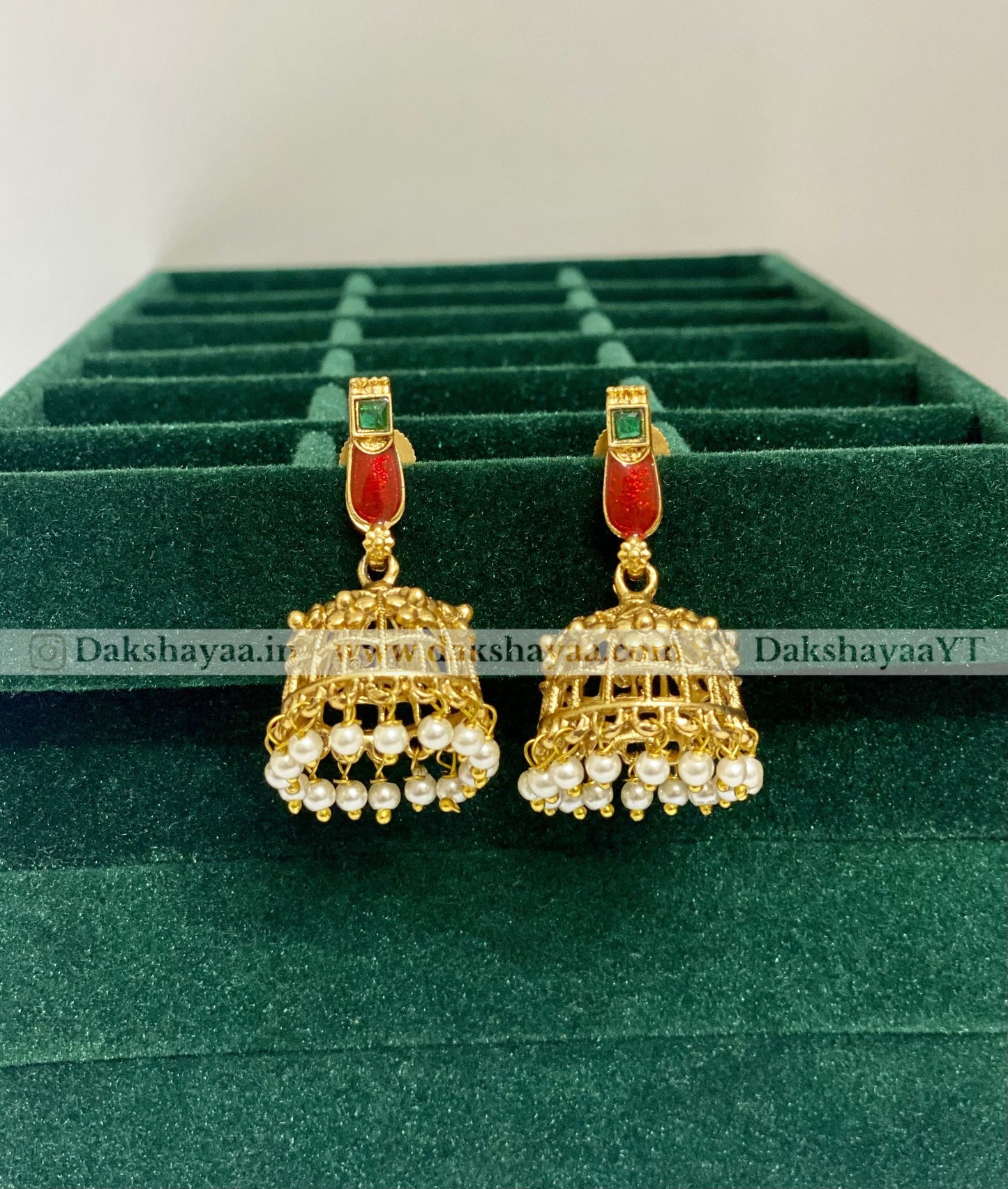Gold and red earrings on a green velvet stand with 'Dakshayaa YT' branding.