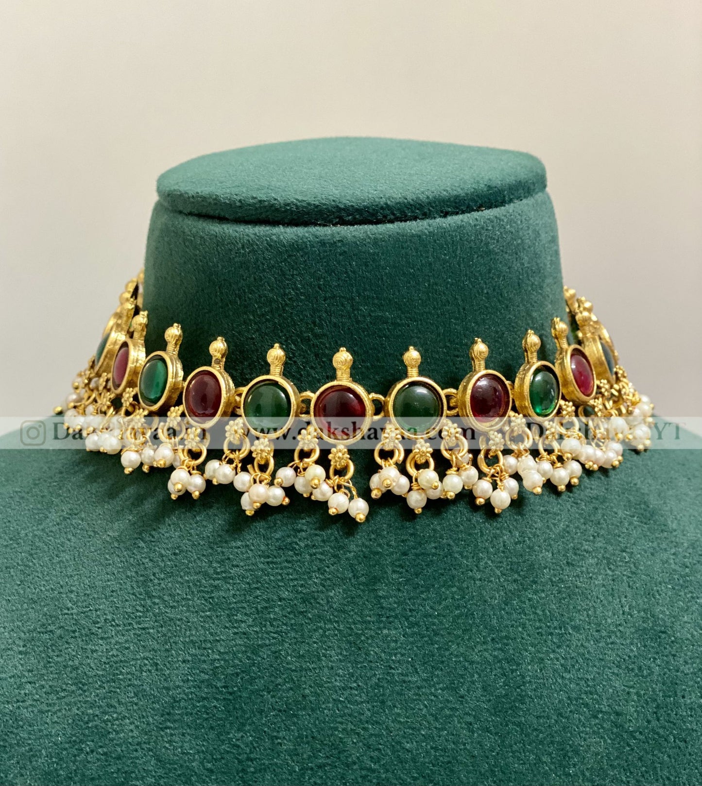 Decorative necklace with gold, red, green, and pearl elements on a green velvet stand.