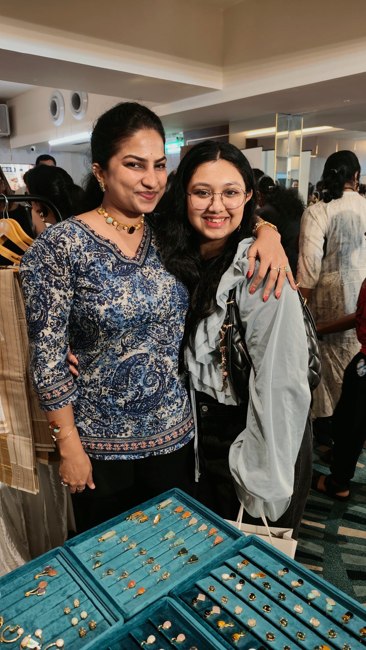 Two women posing together in a jewelry store, with one wearing a blue and white patterned top and the other in a light gray jacket. A teal jewelry display case filled with rings is visible in the foreground.